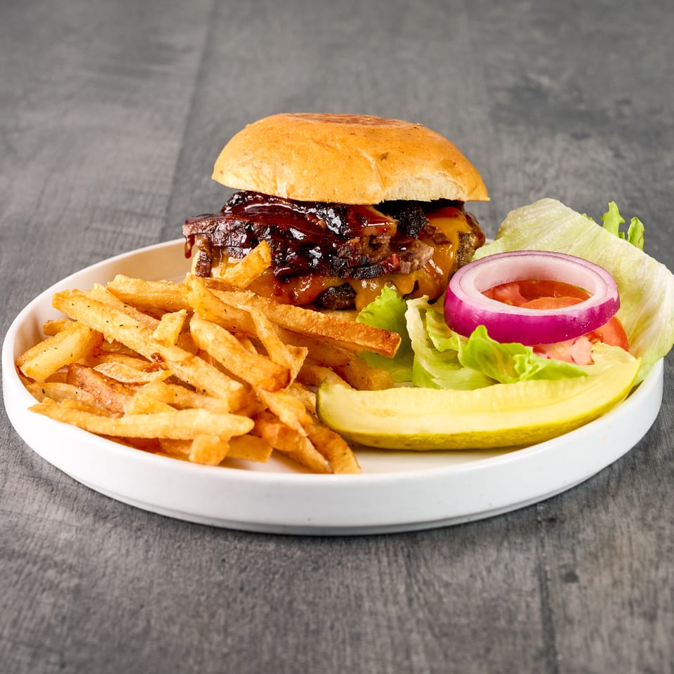A BBQ brisket burger with fries, lettuce, tomato, red onion, and a pickle spear on a white plate.