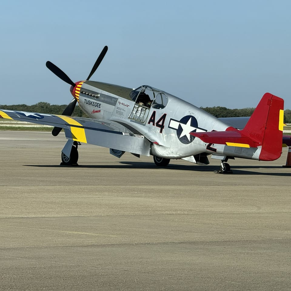Vintage silver-and-red propeller airplane marked “Tuskegee Airmen” parked on an airport runway between orange traffic cones.