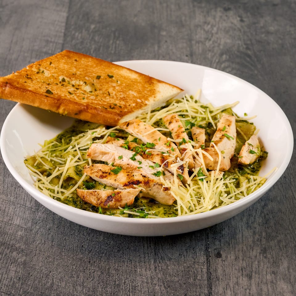 Grilled chicken pesto pasta with shredded cheese and a side of garlic bread in a white bowl.