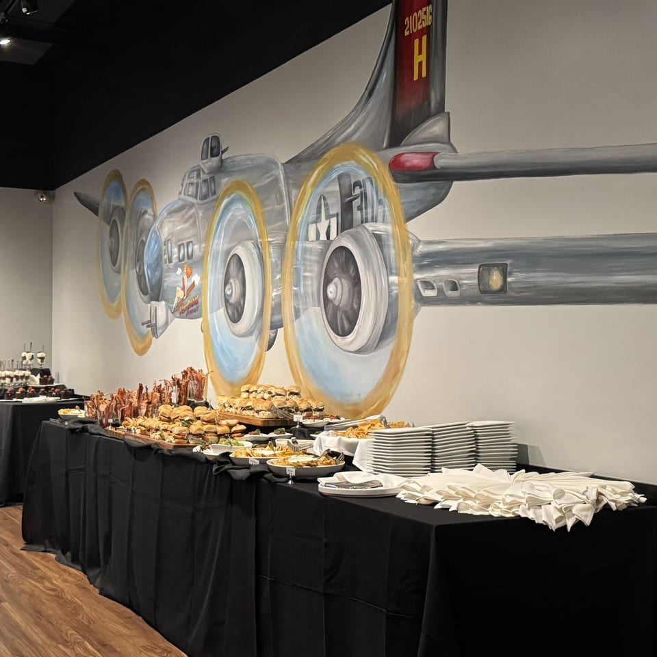 Buffet tables display assorted appetizers, sandwiches, desserts, and stacked plates beneath a large airplane mural.
