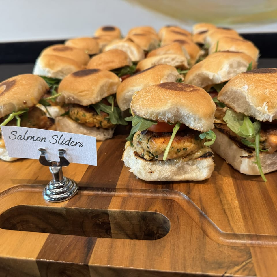 Tray of salmon sliders on toasted buns with greens and tomatoes, labeled “Salmon Sliders.”