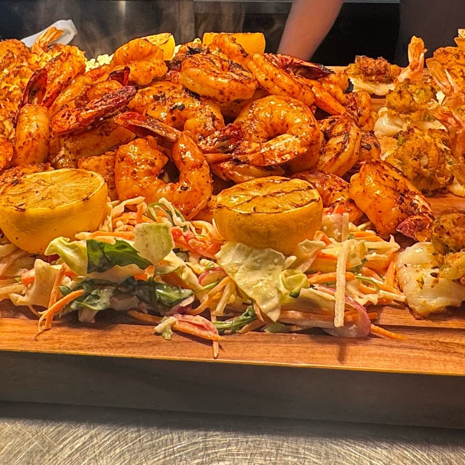 A wooden platter piled with grilled shrimp, crab-stuffed seafood, roasted lemons, and coleslaw.